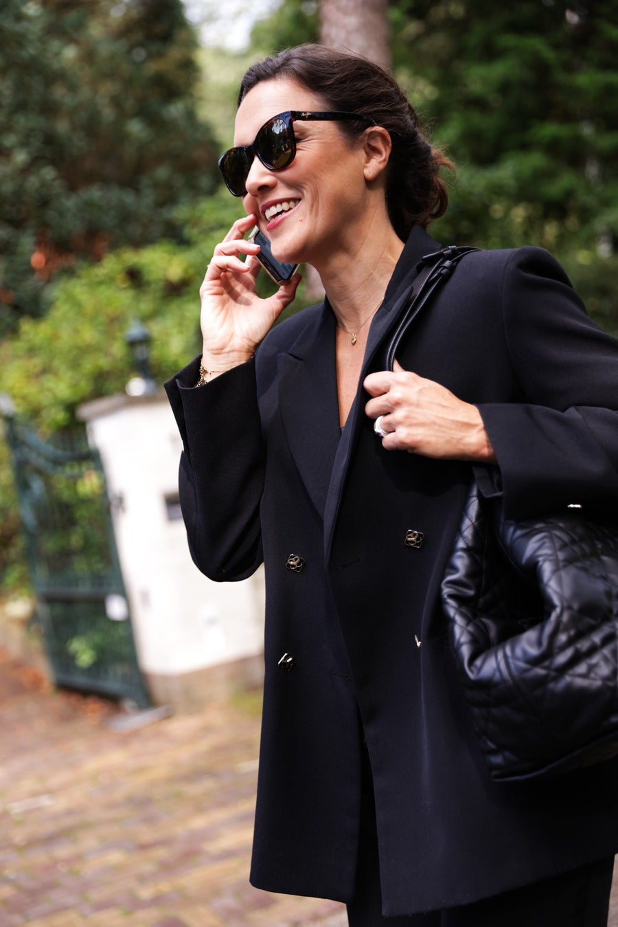 A woman wearing a black double-breasted blazer, black sunglasses, and carrying a black quilted shoulder bag, smiling while talking on a phone in an outdoor garden setting.