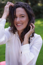 daily wellbeing ritual . A smiling woman with dark wavy hair wearing a white long-sleeve blouse, posing outdoors in a grassy garden setting.