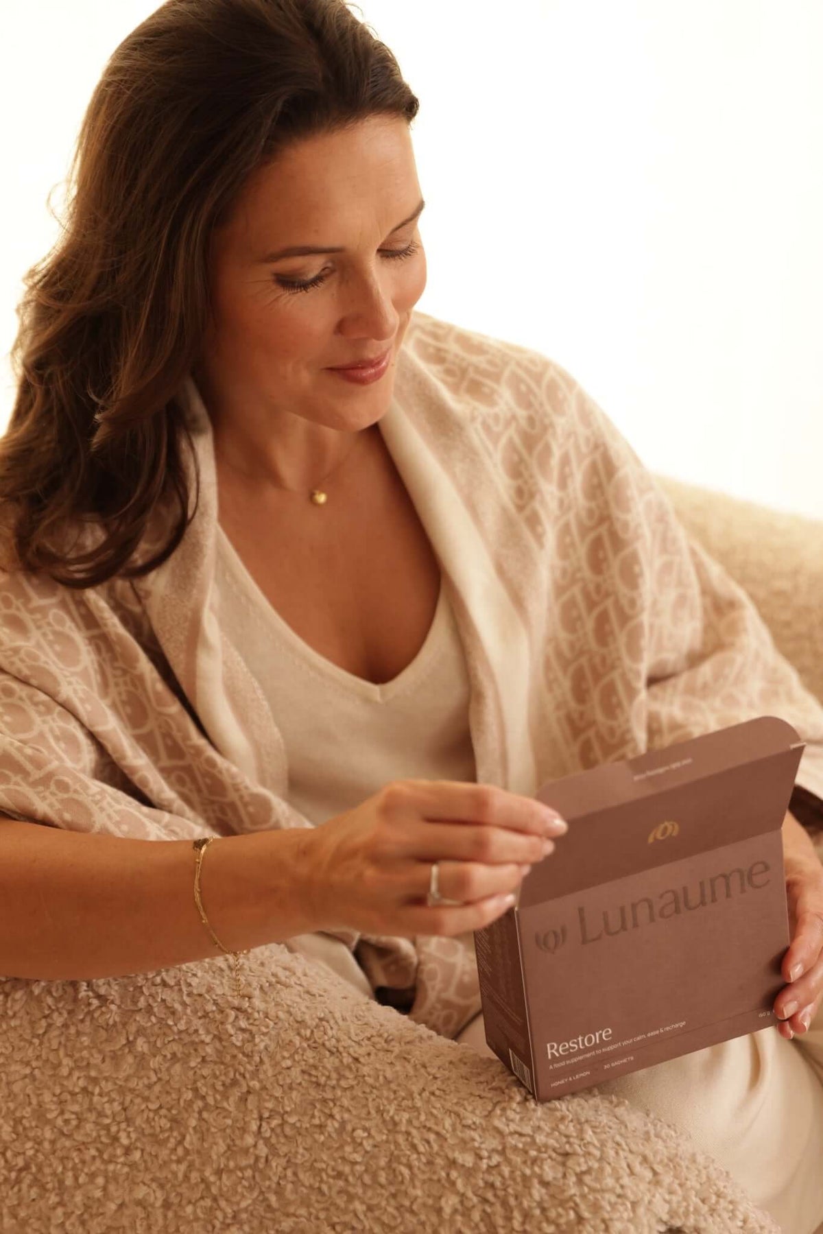 deep recovery sleep supplement. A woman in a beige patterned shawl and cream top sits on a textured couch, holding a brown box labeled 'Lunaume' and 'Restore'.