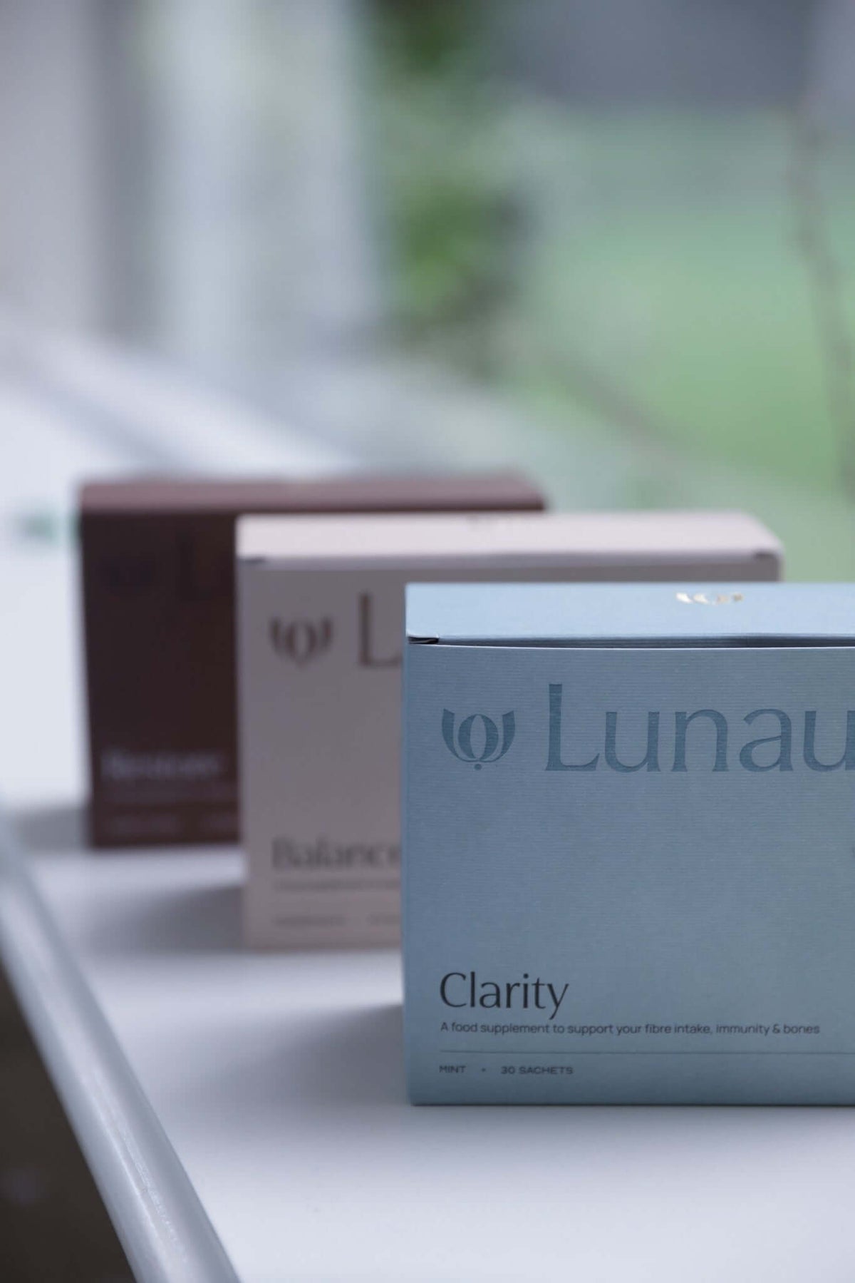 daily wellbeing ritual. A close-up of a Lunau Clarity supplement box in light blue, with two additional Lunau boxes in brown and beige blurred in the background on a white surface near a window.