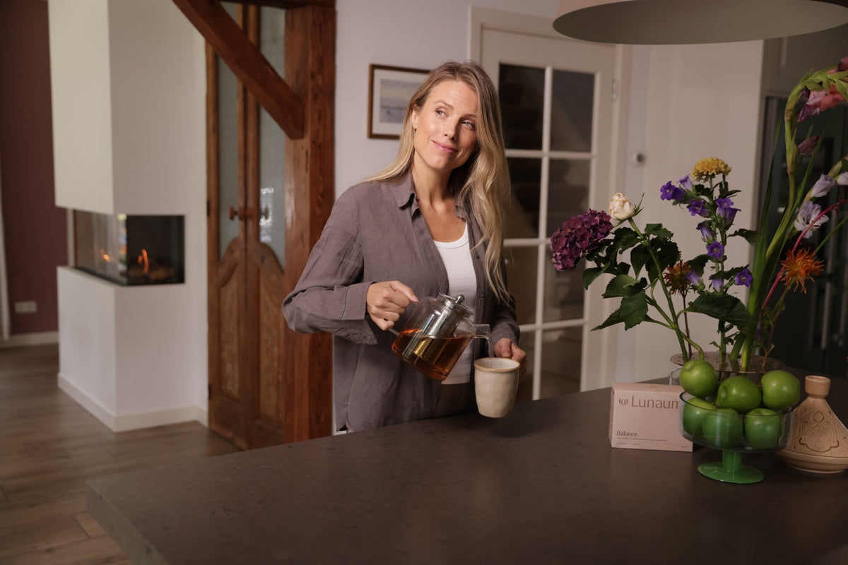 A woman in a modern kitchen setting pours tea from a glass teapot into a mug on a dark countertop. A box labeled 'Lanahta' is visible next to a vase of flowers and green apples.