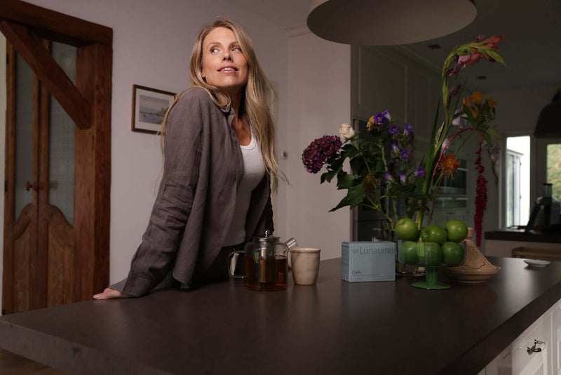 Morning-supplement. A woman with blonde hair leans on a kitchen island, looking upward. The scene includes a wooden door, a teapot, a cup, and a vase of flowers on the dark countertop.