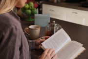 Morning-supplement. A person in a gray shirt reads an open book on a dark kitchen counter, with a mug, glass teapot, and green apples in the background.