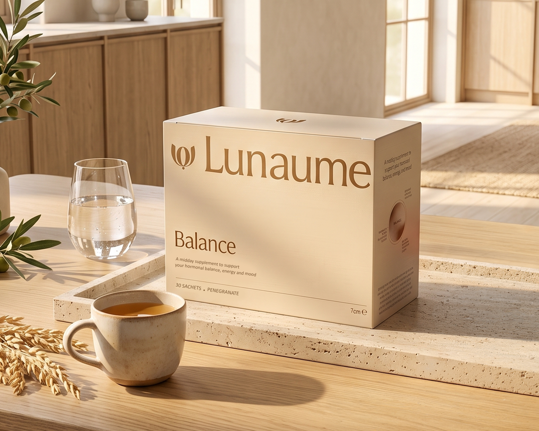 A beige Lunaume Balance supplement box sits on a light stone countertop in a warm, sunlit kitchen setting, with a ceramic cup of tea, a glass of water, and dried wheat stalks nearby.