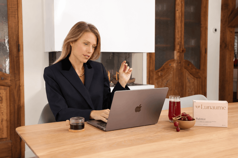 hormonal balance supplement for women. A woman in a black blazer sits at a wooden table using a laptop, with a glass of red juice, a bowl of grapes, and a box of Lunaume in front of her in a modern indoor setting.