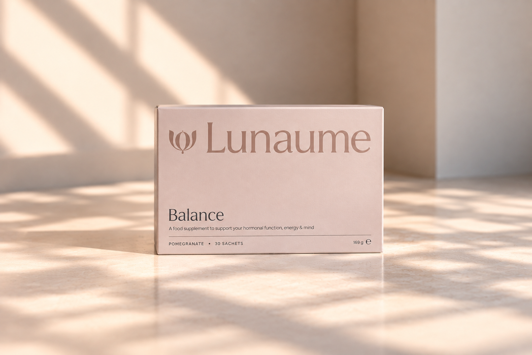 A pale blush pink box of Lunaume Balance dietary supplement, standing upright on a marble surface with warm sunlight and shadow patterns.
