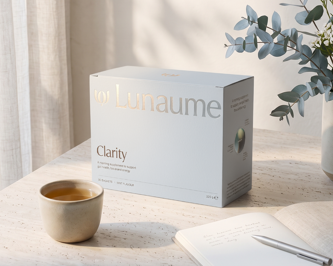 A white box of Lunaume Clarity tea capsules sits on a light stone tabletop beside a small cup of tea, an open notebook, and a pen, with soft sunlight and eucalyptus leaves in the background.