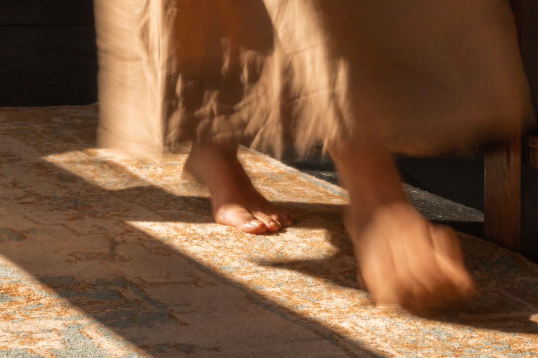A close-up lifestyle shot of a person's bare feet stepping onto a patterned area rug with dramatic, warm sunlight casting shadows across the floor.