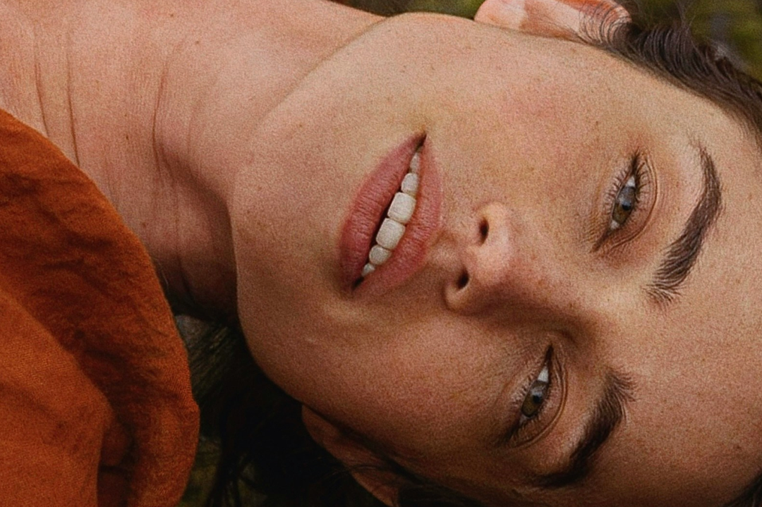Close-up of a woman lying down, looking toward the camera, with a rust-orange textured garment visible on the left side.