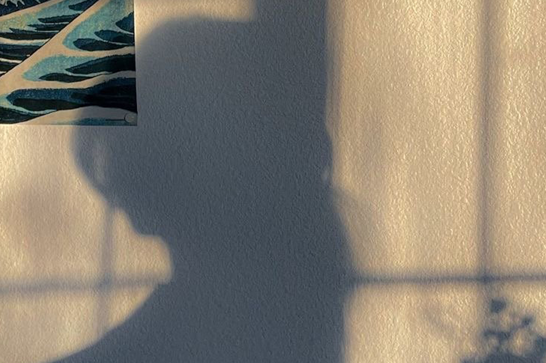 A square art print featuring a textured beige background with a large blue shadow of a person holding a camera, alongside a partial abstract patterned illustration in the top-left corner.