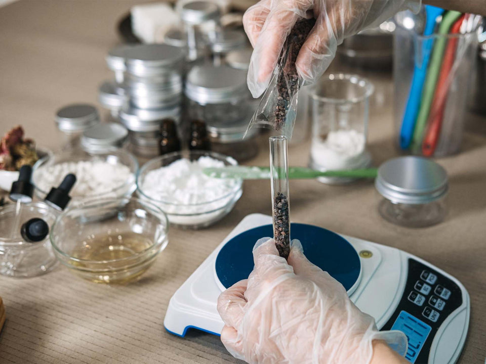 A close-up of gloved hands weighing dark brown or black loose-leaf herbal tea or spices in a clear plastic bag over a digital kitchen scale, with glass jars and bowls of ingredients on a wooden table in the background.