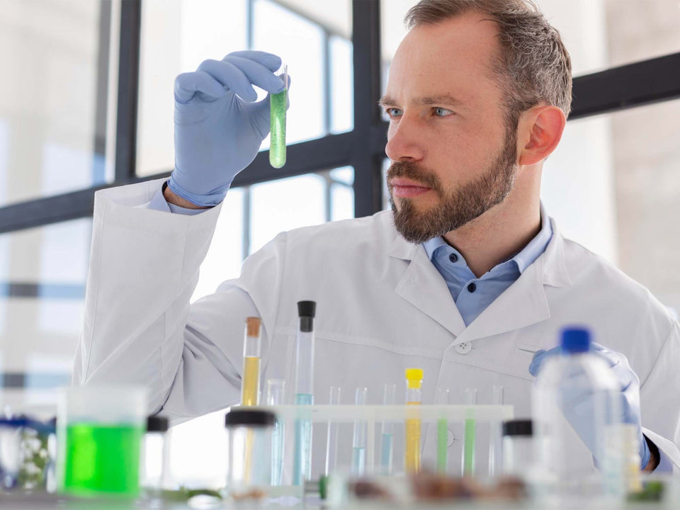 A man in a white lab coat and blue gloves examines a small green test tube held up in a bright laboratory setting with other glassware in the foreground.