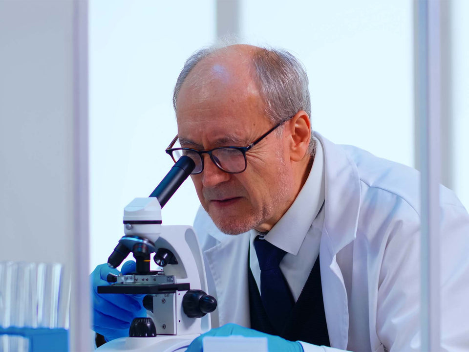 A man in a white lab coat, dark tie, and blue gloves looks through a microscope in a bright laboratory setting.