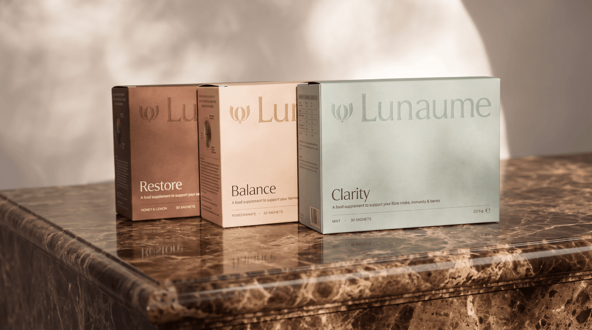 daily wellbeing ritual. Three Lunahme supplement boxes standing on a polished marble surface against a softly lit neutral background. The boxes are labeled Restore, Balance, and Clarity in warm brown, blush, and pale sage tones.